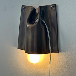 May include: A wall-mounted lamp featuring a dark, smooth, folded ceramic shade with two small holes at the top for mounting. A single glowing light bulb is positioned within the center of the shade, and a clear electrical cord hangs downward.