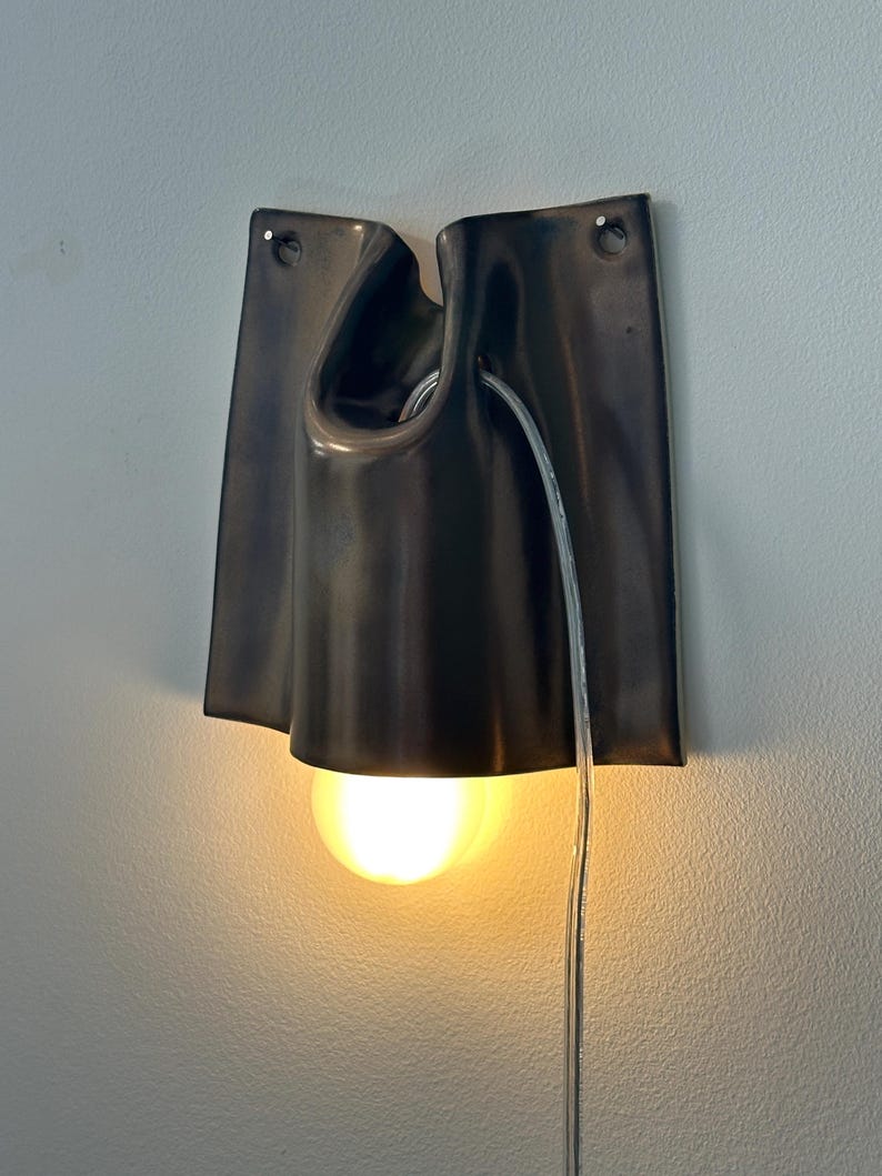 May include: A wall-mounted lamp featuring a dark, smooth, folded ceramic shade with two small holes at the top for mounting. A single glowing light bulb is positioned within the center of the shade, and a clear electrical cord hangs downward.