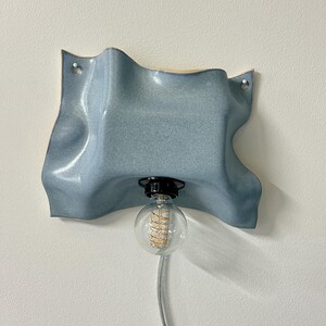 May include: A wall-mounted light fixture featuring a wavy, light blue ceramic shade with a glossy finish. The shade has two small holes for mounting and a central black socket holding a clear, round glass light bulb with a visible spiral filament.
