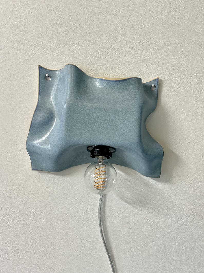 May include: A wall-mounted light fixture featuring a wavy, light blue ceramic shade with a glossy finish. The shade has two small holes for mounting and a central black socket holding a clear, round glass light bulb with a visible spiral filament.