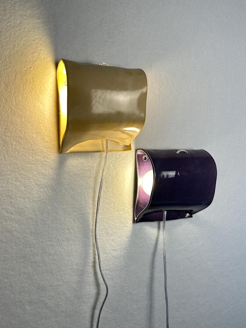 May include: Two wall-mounted sconces with curved, glossy ceramic shades. One shade is light tan and the other is deep purple. Each fixture features a single exposed light bulb emitting a warm glow, with a clear power cord hanging downward.