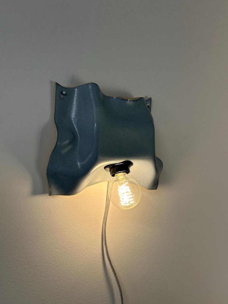 May include: A wall-mounted light fixture featuring a blue, wavy, sheet-like shade with a textured surface. A black socket holds a single, clear, round Edison-style light bulb that emits a warm glow. A thin, light-colored power cord hangs downward.