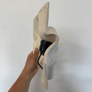 May include: A wall-mounted light fixture with a wavy, off-white ceramic shade featuring black speckles. The fixture includes a black socket and a white and black electrical cord extending from the back of the curved, textured material.