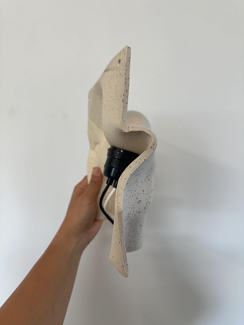 May include: A wall-mounted light fixture with a wavy, off-white ceramic shade featuring black speckles. The fixture includes a black socket and a white and black electrical cord extending from the back of the curved, textured material.