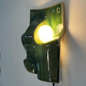 May include: A wall-mounted ceramic sconce featuring a glossy, deep green glazed finish with an organic, undulating shape. A single, spherical, warm-toned light bulb is nestled within a central cutout, casting a soft glow against the wall surface.
