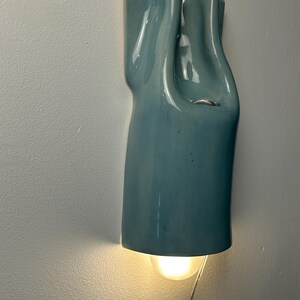 May include: A wall-mounted light fixture with a glossy, light blue ceramic shade featuring a fluid, draped silhouette. The fixture emits a warm, soft glow from a bulb positioned at the bottom, with a thin silver cord extending downward.
