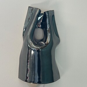 May include: A wall-mounted lamp featuring a glossy, dark charcoal-coloured ceramic shade with an organic, asymmetrical shape. The fixture has a central cutout and a rounded opening at the bottom, revealing a light bulb and a clear power cord.