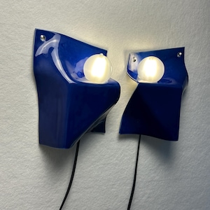 May include: A pair of wall-mounted lamps featuring glossy, deep blue, sculptural ceramic shades with irregular, folded edges. Each lamp includes a single, spherical, frosted white glass bulb that emits a warm glow, connected by a black cord.