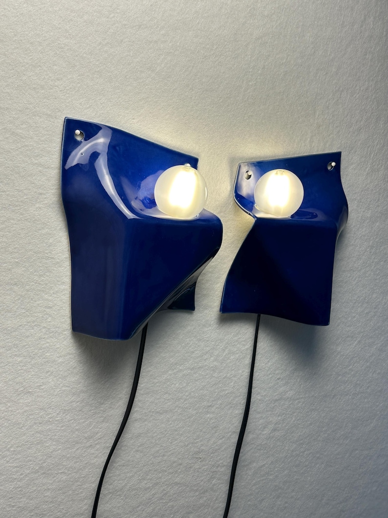 May include: A pair of wall-mounted lamps featuring glossy, deep blue, sculptural ceramic shades with irregular, folded edges. Each lamp includes a single, spherical, frosted white glass bulb that emits a warm glow, connected by a black cord.