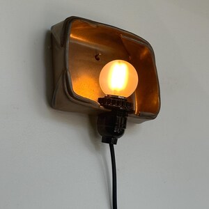 May include: A vintage-style wall sconce with a warm, glowing light bulb. The sconce has a metallic, rectangular shade with a textured surface. A black cord extends from the light fixture.