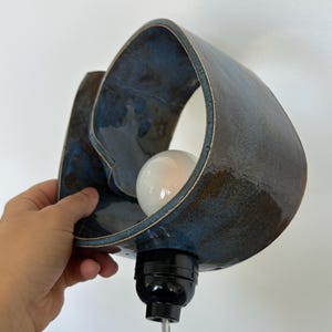 May include: A ceramic wall sconce with a blue and brown glaze. The sconce has a unique, organic shape with a bulb inside. The fixture is held by a hand against a white wall.
