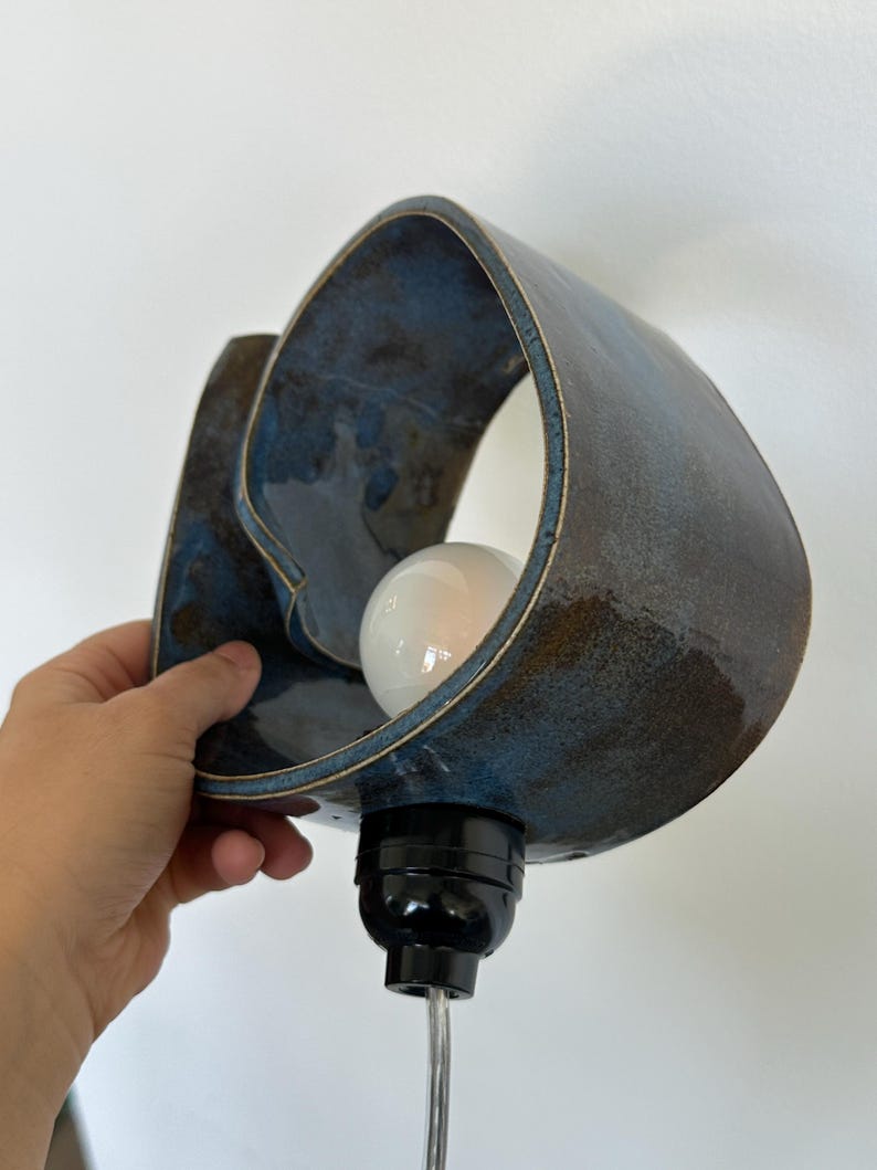 May include: A ceramic wall sconce with a blue and brown glaze. The sconce has a unique, organic shape with a bulb inside. The fixture is held by a hand against a white wall.