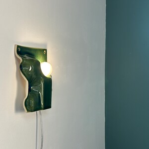 May include: A wall-mounted ceramic light fixture featuring a glossy, deep green, organic, undulating surface. The piece has a rectangular backplate with two visible mounting holes and a circular, warm-glowing light bulb protruding from the side.