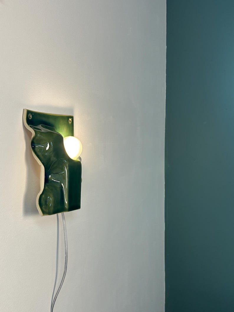 May include: A wall-mounted ceramic light fixture featuring a glossy, deep green, organic, undulating surface. The piece has a rectangular backplate with two visible mounting holes and a circular, warm-glowing light bulb protruding from the side.