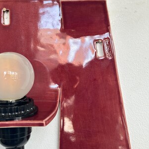 以下が含まれることがあります： 光沢のある仕上がりの、バーガンディ色の壁掛け照明。電球と黒いベースが付いた、湾曲した棚が特徴です。「T-MC00D」の文字が入った切り欠きのある長方形のデザインです。