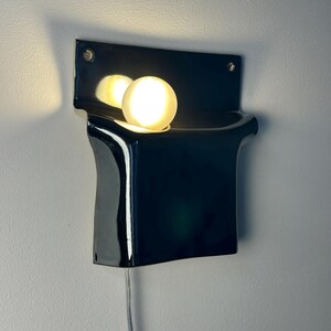 Peut inclure: Une applique murale compos&eacute;e d'une base en c&eacute;ramique noire brillante aux formes courbes et sculpturales. Une ampoule ronde en verre d&eacute;poli blanc est log&eacute;e dans la partie sup&eacute;rieure du luminaire, diffusant une lueur chaude sur la surface.