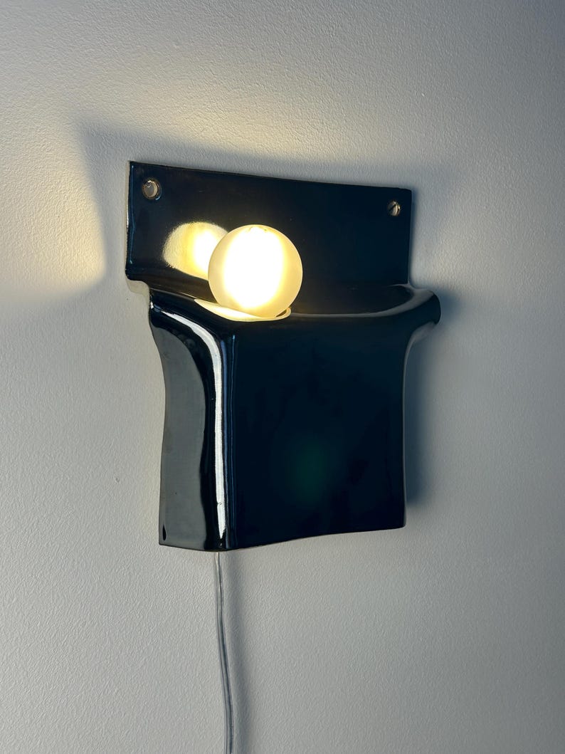 Peut inclure: Une applique murale compos&eacute;e d'une base en c&eacute;ramique noire brillante aux formes courbes et sculpturales. Une ampoule ronde en verre d&eacute;poli blanc est log&eacute;e dans la partie sup&eacute;rieure du luminaire, diffusant une lueur chaude sur la surface.