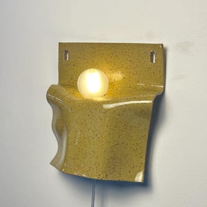 May include: A wall-mounted ceramic lamp with a speckled mustard yellow glaze and a wavy, folded form. The fixture features a central circular opening holding a glowing, frosted white spherical light bulb and includes two rectangular mounting holes.