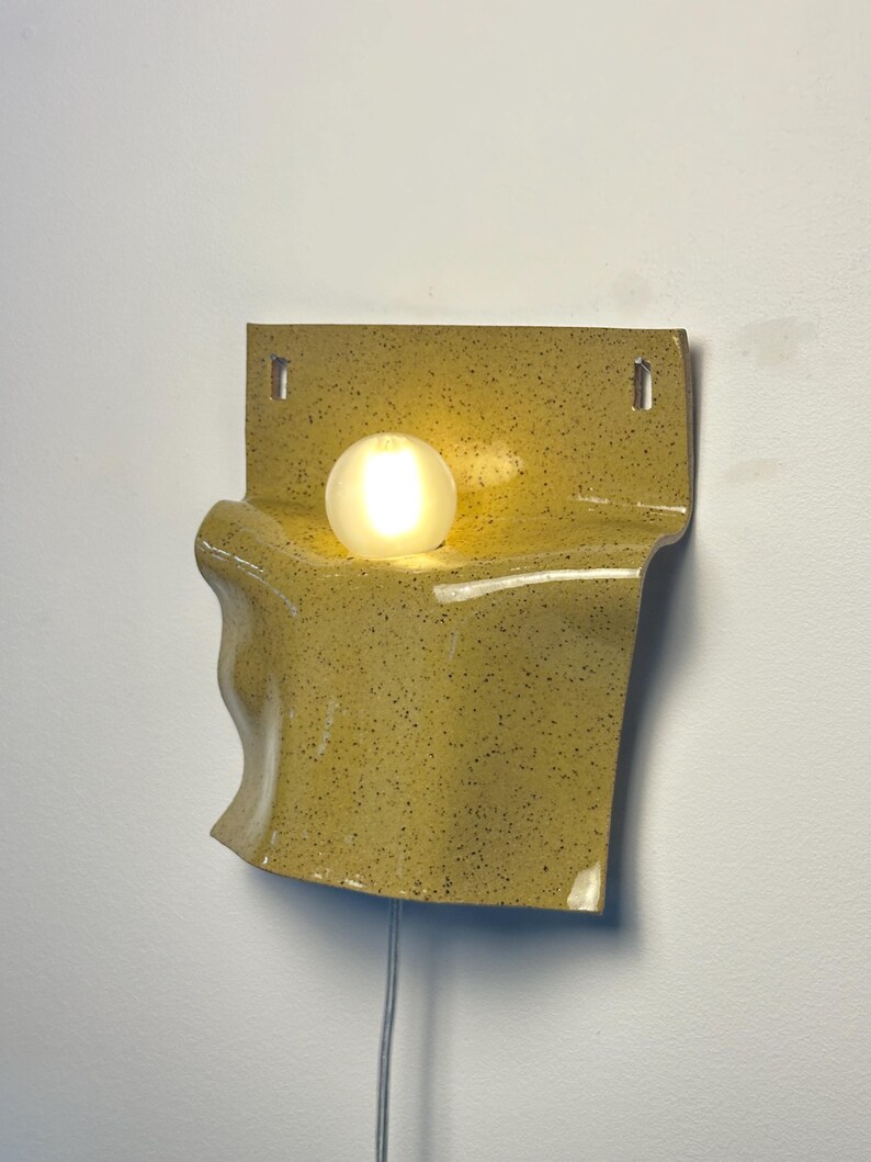 May include: A wall-mounted ceramic lamp with a speckled mustard yellow glaze and a wavy, folded form. The fixture features a central circular opening holding a glowing, frosted white spherical light bulb and includes two rectangular mounting holes.