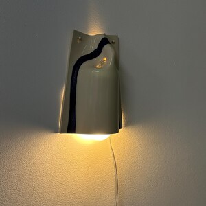 May include: A wall-mounted ceramic light fixture with a glossy off-white finish and a single dark blue vertical stripe. The curved, folded form emits a warm glow from the top and bottom, and a clear power cord hangs from the base of the unit.