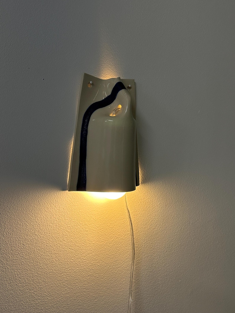 May include: A wall-mounted ceramic light fixture with a glossy off-white finish and a single dark blue vertical stripe. The curved, folded form emits a warm glow from the top and bottom, and a clear power cord hangs from the base of the unit.