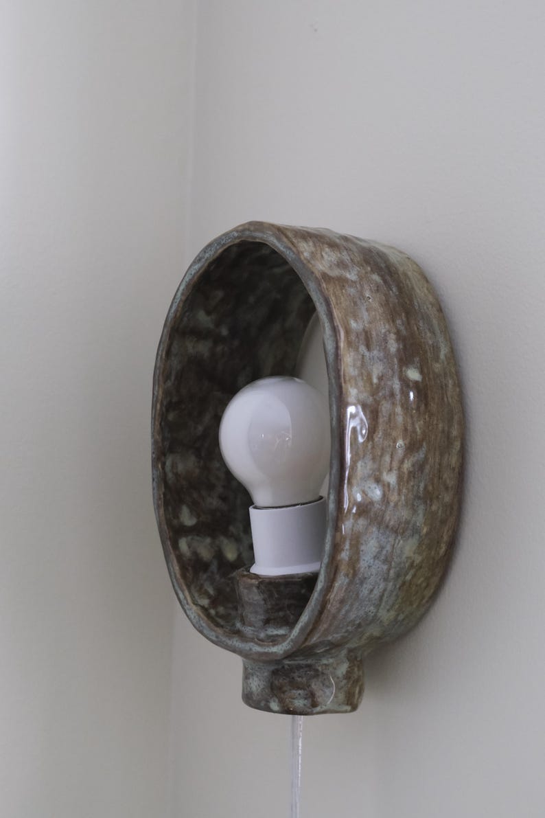 May include: A ceramic wall sconce with a bulb. The sconce has a brown and blue-green mottled glaze. The light bulb is white. The sconce is mounted on a white wall. The fixture is a decorative lighting element.