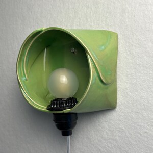 May include: A wall-mounted light fixture featuring a curved, light green ceramic shade with a glossy finish. A black socket holds a frosted, spherical light bulb inside the ceramic housing, with a thin gray power cord extending downward.