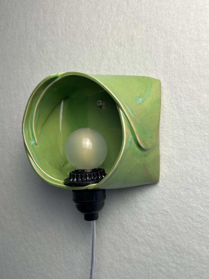 May include: A wall-mounted light fixture featuring a curved, light green ceramic shade with a glossy finish. A black socket holds a frosted, spherical light bulb inside the ceramic housing, with a thin gray power cord extending downward.