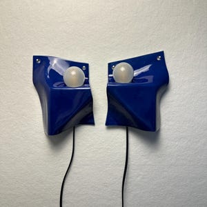 May include: A pair of glossy, deep blue wall-mounted lamps with an asymmetrical, folded geometric shape. Each lamp features a single, spherical, frosted white light bulb positioned in a recessed area, with a thin black power cord extending downward.