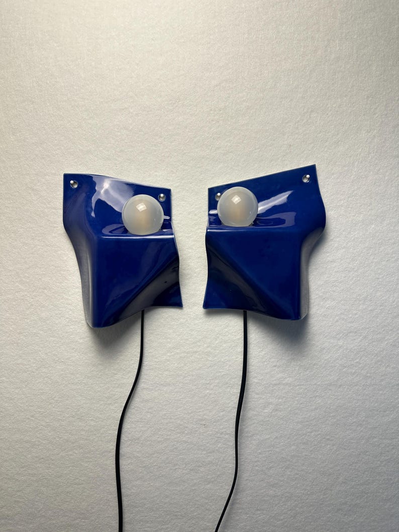 May include: A pair of glossy, deep blue wall-mounted lamps with an asymmetrical, folded geometric shape. Each lamp features a single, spherical, frosted white light bulb positioned in a recessed area, with a thin black power cord extending downward.
