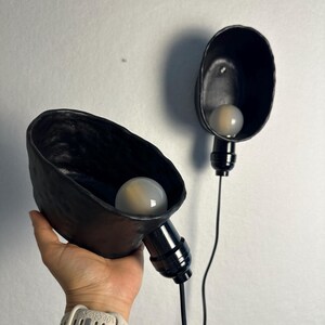 K&ouml;nnte beinhalten: Zwei Wandleuchten mit unregelm&auml;&szlig;ig geformten, handgefertigten schwarzen Keramikschirmen in matter Optik. Jede Leuchte verf&uuml;gt &uuml;ber eine schwarze Fassung und eine runde, wei&szlig; mattierte Gl&uuml;hbirne. Die Schirme haben eine organische Form.