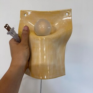 May include: A wall-mounted ceramic lamp with a glossy, pale yellow glaze. The fixture features a curved, concave shape with two circular holes at the top for mounting. A single clear glass light bulb is centered in the middle, and a white electrical cord with a two-prong plug is attached.
