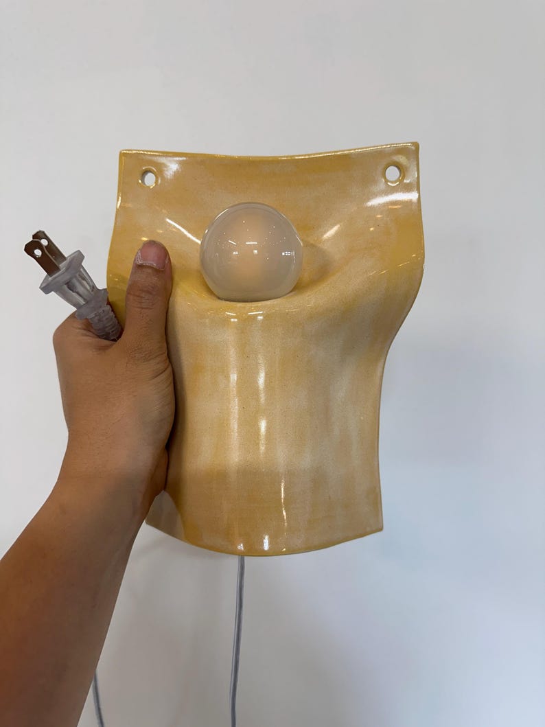 May include: A wall-mounted ceramic lamp with a glossy, pale yellow glaze. The fixture features a curved, concave shape with two circular holes at the top for mounting. A single clear glass light bulb is centered in the middle, and a white electrical cord with a two-prong plug is attached.