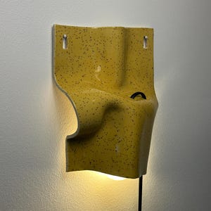 K&ouml;nnte beinhalten: Eine gelbe Wandleuchte mit schwarzem Sprenkelmuster und einer geschwungenen, gefalteten Keramikform. Die Leuchte verf&uuml;gt &uuml;ber zwei obere Befestigungsl&ouml;cher, eine mittige schwarze Kabelschlaufe und eine nach unten gerichtete Lichtquelle.