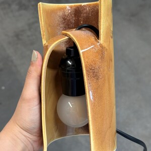 Puede incluir: Una l&aacute;mpara cil&iacute;ndrica de cer&aacute;mica con un esmalte brillante moteado en tonos &aacute;mbar y marr&oacute;n. La estructura presenta un dise&ntilde;o curvo abierto en la parte frontal con un casquillo negro y una bombilla esf&eacute;rica blanca suspendida por un cable negro.