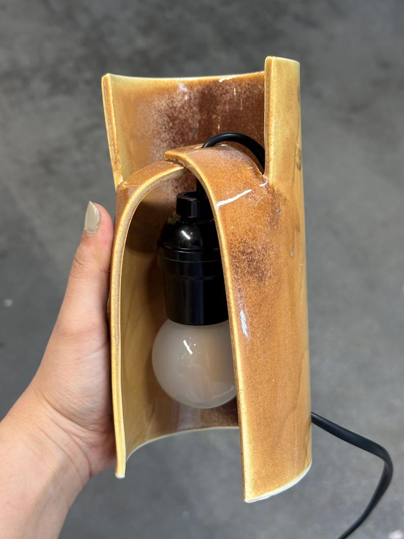 Puede incluir: Una l&aacute;mpara cil&iacute;ndrica de cer&aacute;mica con un esmalte brillante moteado en tonos &aacute;mbar y marr&oacute;n. La estructura presenta un dise&ntilde;o curvo abierto en la parte frontal con un casquillo negro y una bombilla esf&eacute;rica blanca suspendida por un cable negro.