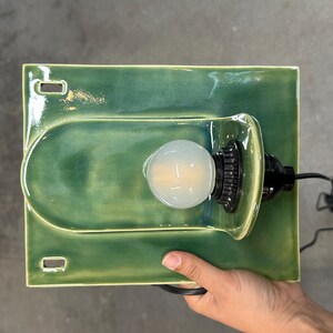 May include: A rectangular green ceramic wall sconce featuring a glossy finish and a central arched protrusion. The fixture includes a black socket holding a round, frosted light bulb and a black electrical cord extending from the side.