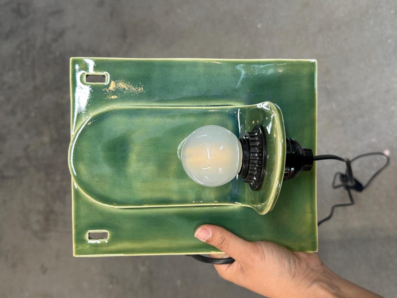May include: A rectangular green ceramic wall sconce featuring a glossy finish and a central arched protrusion. The fixture includes a black socket holding a round, frosted light bulb and a black electrical cord extending from the side.