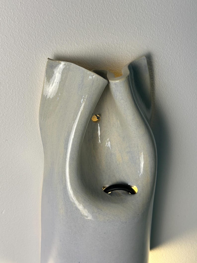 May include: A pale blue ceramic wall sconce with a smooth, undulating, organic form. The fixture features a glossy finish and two small, circular cutouts that reveal a warm, glowing light source from within the hollow, curved structure.