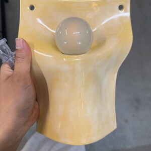 May include: A wall-mounted light fixture featuring a curved, pale yellow ceramic shade with a glossy finish. The shade has two circular mounting holes at the top corners and a central opening that holds a single, round, frosted white glass light bulb.