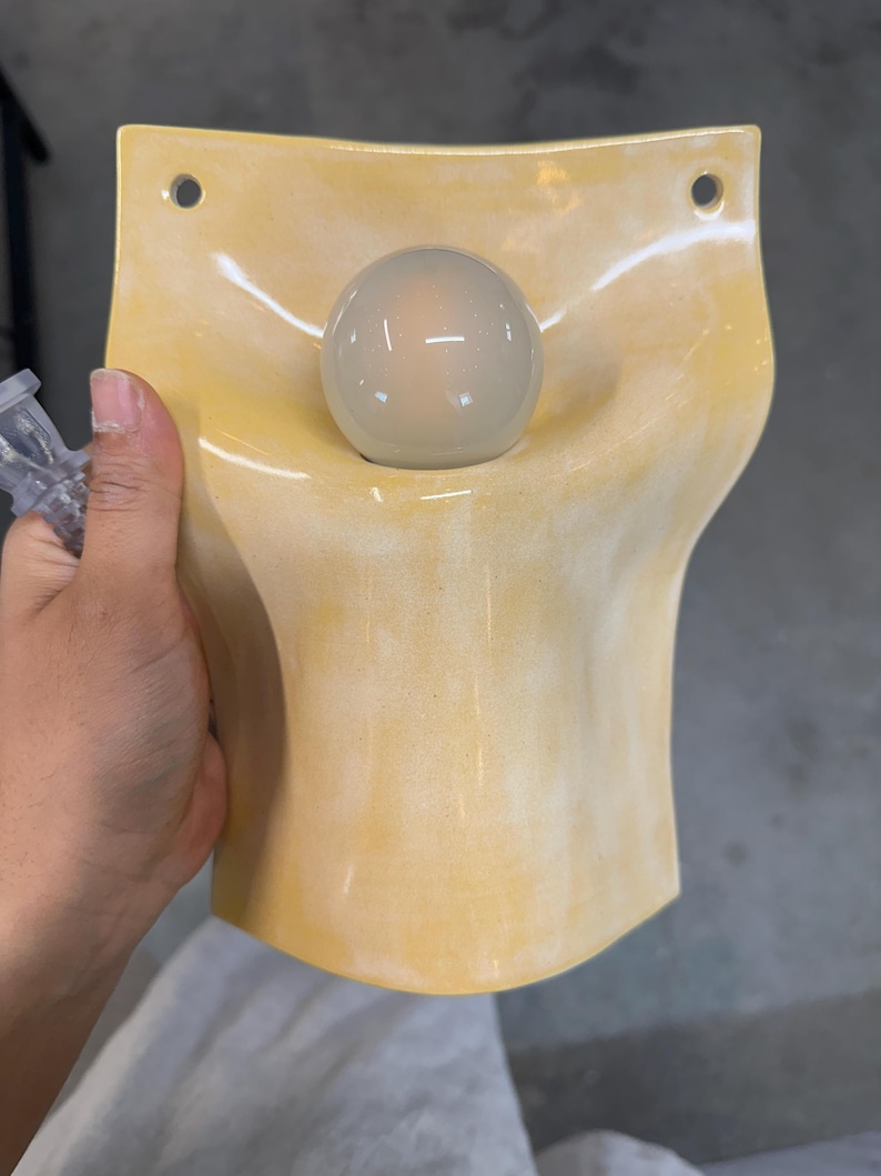 May include: A wall-mounted light fixture featuring a curved, pale yellow ceramic shade with a glossy finish. The shade has two circular mounting holes at the top corners and a central opening that holds a single, round, frosted white glass light bulb.