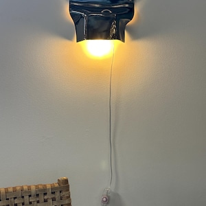 May include: A glossy dark blue wall-mounted lamp with a curved, flared shade design. The fixture emits a warm, bright yellow light downward. A thin white power cord extends from the base, featuring a small inline switch for operation.