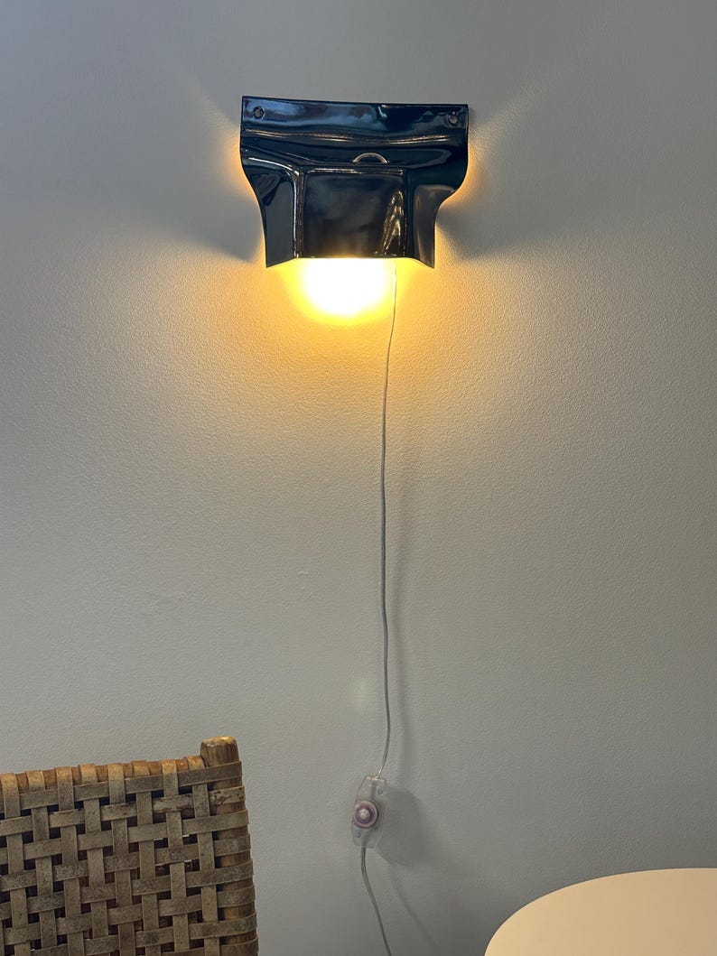 May include: A glossy dark blue wall-mounted lamp with a curved, flared shade design. The fixture emits a warm, bright yellow light downward. A thin white power cord extends from the base, featuring a small inline switch for operation.