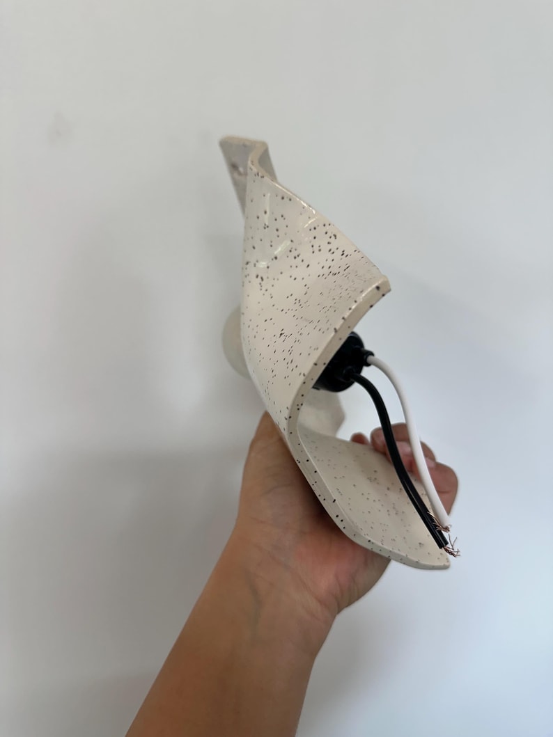 Może przedstawiać: Kinkiet z zakrzywionym, kremowym kloszem ceramicznym w czarne kropki. Oprawa zawiera czarną oprawkę żar&oacute;wki oraz dwa odsłonięte przewody elektryczne, czarny i biały, wystające ze środka elementu oświetleniowego w kierunku zewnętrznym.