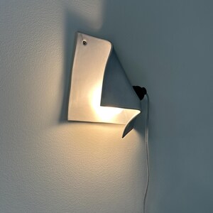May include: A white ceramic wall sconce with a curved, folded sheet design. The fixture features a single glowing light bulb tucked behind the fold, a small mounting hole at the top corner, and a thin white electrical cord extending downward.