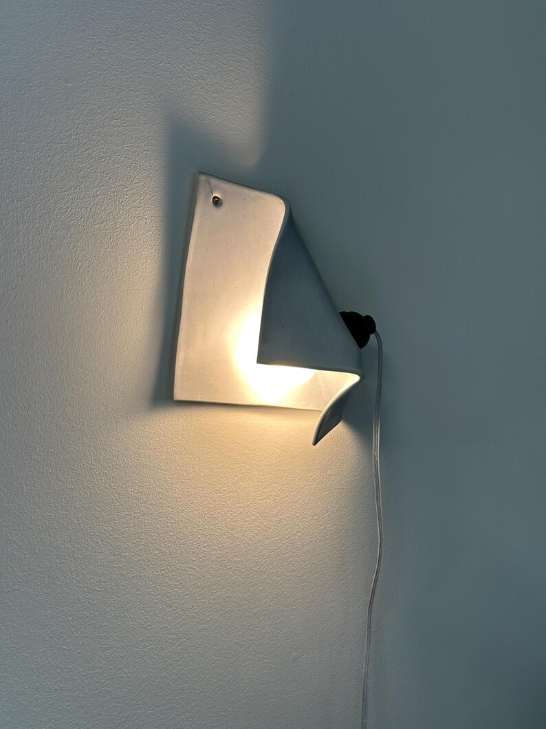 May include: A white ceramic wall sconce with a curved, folded sheet design. The fixture features a single glowing light bulb tucked behind the fold, a small mounting hole at the top corner, and a thin white electrical cord extending downward.