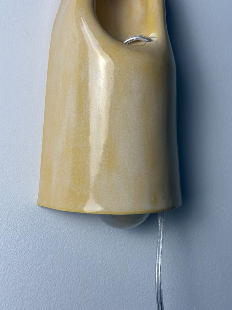 Pu&ograve; includere: Una lampada da parete in ceramica con una finitura lucida color giallo senape. Il paralume a forma di campana presenta un'apertura circolare vicino alla parte superiore, che rivela un anello di metallo argentato all'interno. Un cavo trasparente pende.