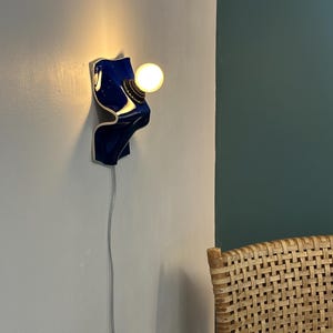 May include: A wall-mounted lamp featuring a glossy, deep blue ceramic base with a curved, wave-like form. The fixture includes a brass-toned socket holding a single, glowing spherical light bulb. A thin, light-colored electrical cord hangs downward.