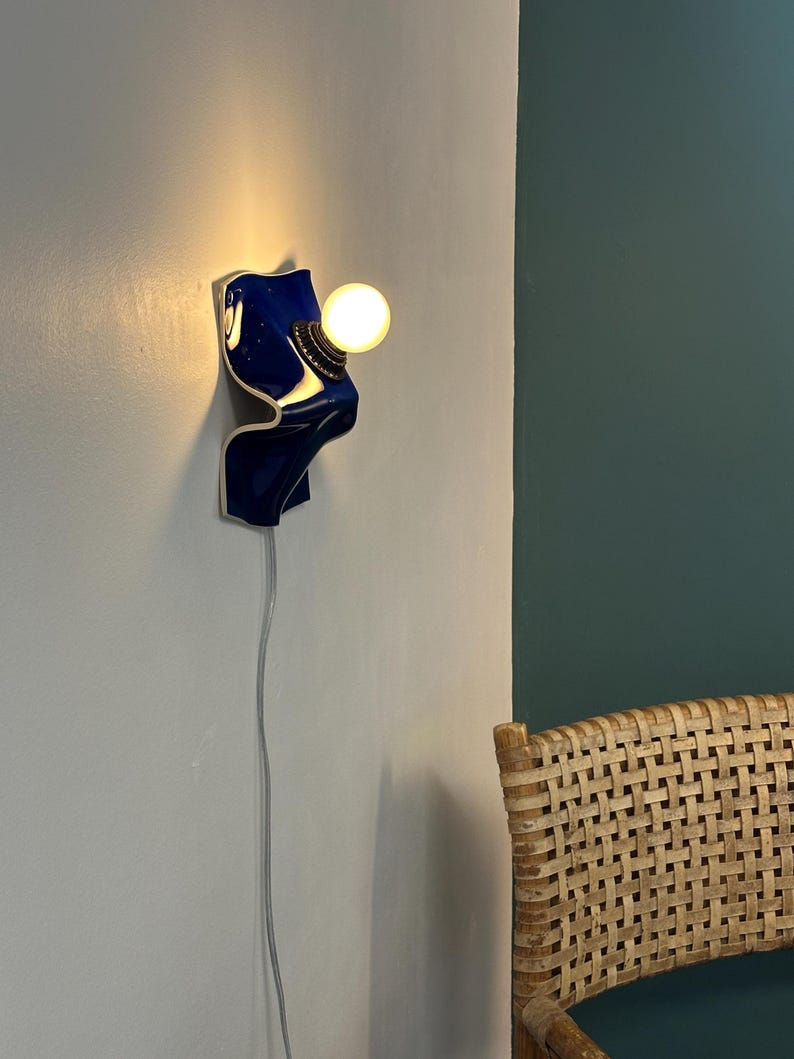 May include: A wall-mounted lamp featuring a glossy, deep blue ceramic base with a curved, wave-like form. The fixture includes a brass-toned socket holding a single, glowing spherical light bulb. A thin, light-colored electrical cord hangs downward.