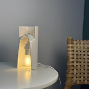 May include: A white ceramic table lamp with a curved, cylindrical structure that features an open front. The lamp houses a single light bulb suspended from the top interior, emitting a warm glow through the opening of the white ceramic body.